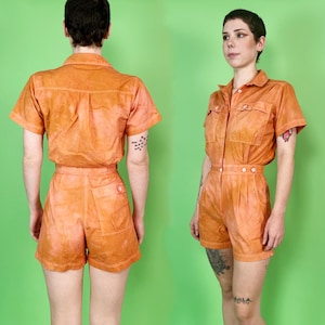 May include: Orange short-sleeved jumpsuit with a collared neckline, two chest pockets, and a button-down front. The shorts have a high waist and a single back pocket. The fabric has a tie-dye effect. The model is standing in front of a green background.