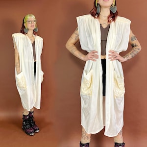 May include: A white sleeveless long vest with a shawl collar and two large pockets with lace trim. The vest is made of a lightweight fabric and has a relaxed fit.