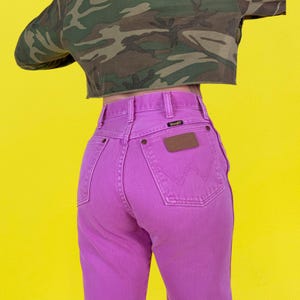May include: A pair of bright pink Wrangler jeans with a brown leather patch and a small black label. The jeans are high-waisted and have a classic five-pocket design. The background is a bright yellow.