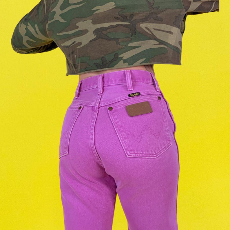 May include: A pair of bright pink Wrangler jeans with a brown leather patch and a small black label. The jeans are high-waisted and have a classic five-pocket design. The background is a bright yellow.