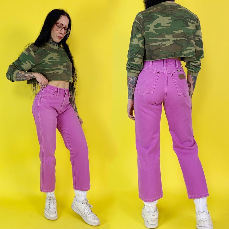May include: A pair of bright pink denim jeans with a high waist and straight leg. The jeans are paired with a cropped camouflage print long-sleeve shirt. The background is a solid yellow.