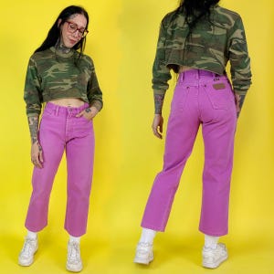 May include: A person wearing a cropped camouflage long-sleeve shirt and high-waisted, straight-leg, magenta jeans. The jeans have a small brown patch on the back pocket. The person is wearing white sneakers and socks. The background is yellow.