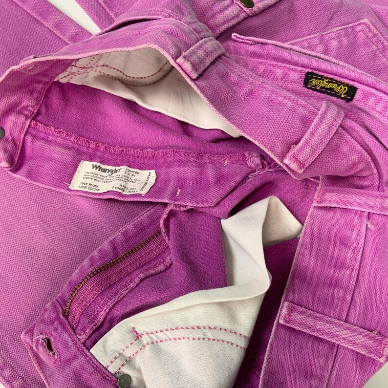 May include: Close-up of a pair of bright pink Wrangler jeans. The jeans are folded, showing the front pockets, zipper, and the Wrangler label. The interior lining is white, and the stitching is visible.