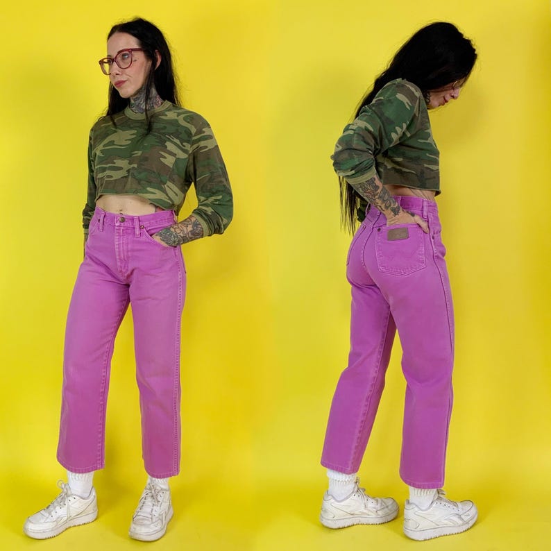May include: A cropped camouflage long-sleeve shirt and bright pink jeans. The jeans have a classic five-pocket design. The outfit is paired with white sneakers and white socks. The background is a solid yellow.