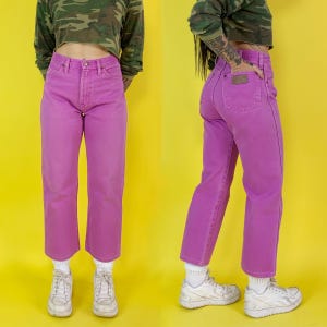 May include: A pair of vibrant, pink denim jeans. The jeans have a classic five-pocket design and a straight leg fit. The jeans are paired with white sneakers and white socks. The background is a bright yellow.