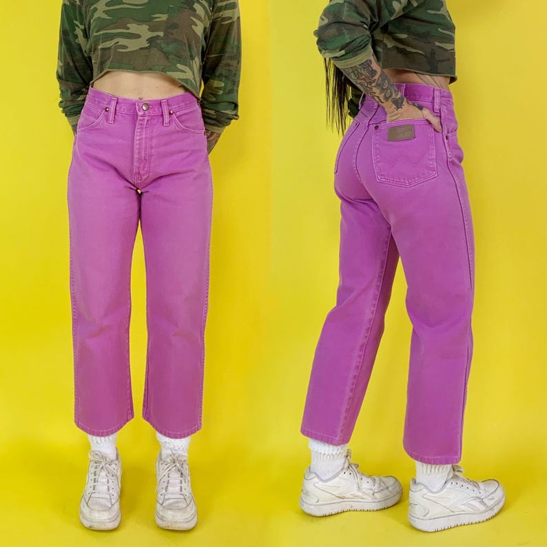 May include: A pair of vibrant, pink denim jeans. The jeans have a classic five-pocket design and a straight leg fit. The jeans are paired with white sneakers and white socks. The background is a bright yellow.