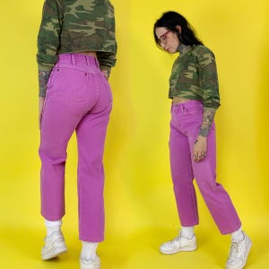 May include: Bright pink denim jeans with a high waist and straight leg. The jeans are paired with a camouflage print long-sleeve crop top. The background is a bright yellow.