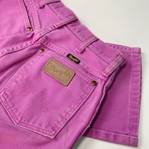 May include: Close-up of a pair of pink Wrangler jeans. The jeans feature a classic five-pocket design, belt loops, and a leather patch with the Wrangler logo. The stitching and rivets are visible, and the jeans are folded.