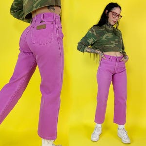 May include: Bright pink denim jeans with a classic five-pocket design and a brown leather patch on the back pocket. The jeans have a straight leg cut and a high waist. The model is wearing a camouflage print top.