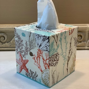 May include: A white tissue box cover with a sea life design. The design features coral, starfish, seashells, and seaweed in shades of red, brown, blue, and green. The cover has a light blue trim.