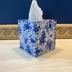 May include: A square tissue box with a white and blue floral pattern. The box is filled with white tissues, with a few tissues extending out of the top. The background is a solid dark blue.
