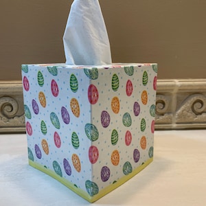 May include: A white tissue box cover with a colorful Easter egg pattern. The cover has a yellow border and is decorated with green, pink, orange, and purple eggs on a white background.
