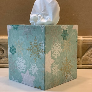 May include: A square tissue box cover with a snowflake pattern in shades of green, white, and gold. The cover is made of fabric and has a white tissue box inside.