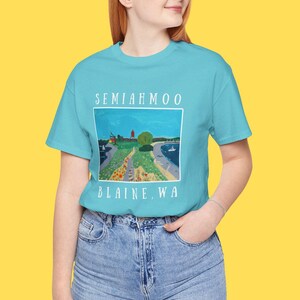 Könnte beinhalten: Ein türkisfarbenes T-Shirt mit einer Illustration der Küstenlandschaft von Semiahmoo, Blaine, WA. Das Kunstwerk zeigt einen Leuchtturm, Bäume und einen Weg. Der Text "SEMIAHMOO" und "BLAINE, WA" sind oberhalb und unterhalb des Bildes aufgedruckt.