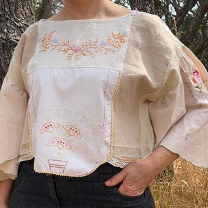 May include: A cream-coloured, embroidered top with bell sleeves. The top features floral embroidery in pink, blue, and orange. The sleeves have a scalloped edge. The top is made of a light, airy fabric.
