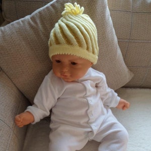 Puede incluir: Una muñeca bebé con un gorro de punto amarillo pálido con un diseño en espiral y un pequeño pompón en la parte superior. La muñeca está vestida con un mono blanco. La muñeca está sentada sobre una superficie blanca.