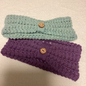 May include: Two knitted headbands with wooden buttons. The headband on top is light green and the headband on the bottom is purple.