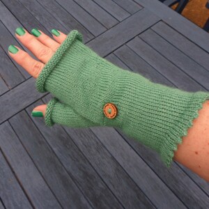 Könnte beinhalten: Grüne fingerlose Handschuhe mit einem Holzknopf. Die Handschuhe sind gestrickt und haben einen gewellten Rand.