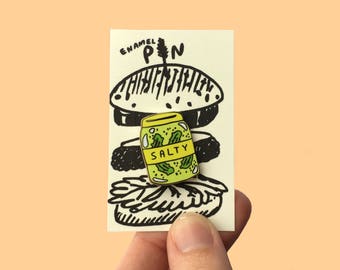 May include: Enamel pin of a yellow jar with green pickles inside. The jar has the word "SALTY" printed on it. The pin is on a white card with a black and white illustration of a burger.