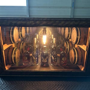 May include: A wooden display cabinet with a dark frame, featuring miniature whiskey barrels and bottles. The interior is lit with a warm light, highlighting the bottles and barrels, creating a rustic atmosphere. The bottles have labels with text.