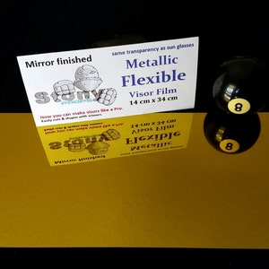 May include: A gold metallic flexible visor film sheet, 14 inches by 34 inches, with a mirror finish. The sheet is laying flat on a black surface. The text "Metallic Flexible Visor Film 14 cm x 34 cm" is printed on a white card behind the sheet.