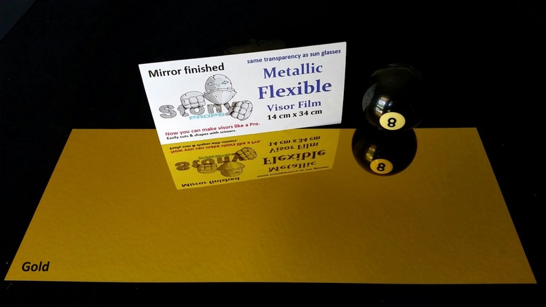May include: A gold metallic flexible visor film sheet, 14 inches by 34 inches, with a mirror finish. The sheet is laying flat on a black surface. The text "Metallic Flexible Visor Film 14 cm x 34 cm" is printed on a white card behind the sheet.