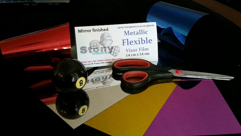 May include: A red, blue, and gold metallic flexible visor film with a mirror finish. The film is 14 inches by 34 inches and comes with a pair of scissors. The packaging includes the text "Mirror finished", "Stony", "same transparency as sun glasses", "Metallic Flexible", "Visor Film", "14 cm x 34 cm", and a cartoon image of a robot.