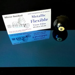 May include: A blue, mirror-finished, flexible visor film sheet with the text "Metallic Flexible Visor Film 14 cm x 34 cm" and a drawing of a person wearing a visor. The sheet is next to a black 8-ball with a yellow number 8.