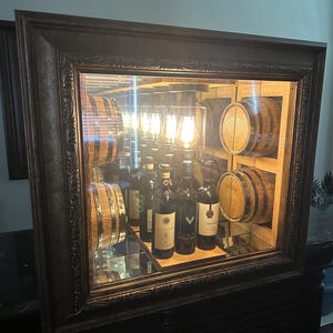 May include: A framed wine cabinet with a dark wood finish. The cabinet features a mirrored interior with wooden barrels and shelves holding wine bottles. Warm lighting illuminates the display, creating a sophisticated ambiance.
