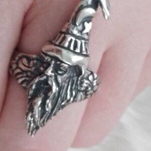 May include: A silver ring featuring a detailed sculpture of a wizard with a pointed hat and a long beard. The wizard has wings on his back and a star on his hat.