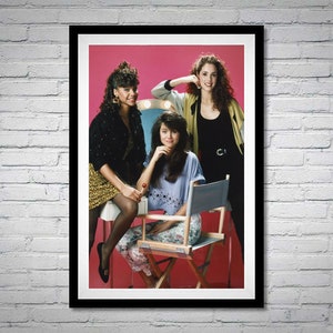 May include: A framed photograph of three women posing in front of a pink background. The woman in the middle is sitting on a director's chair, while the other two women are standing behind her. The woman on the left is wearing a black jacket and a gold skirt, while the woman on the right is wearing a black tank top and a beige cardigan.