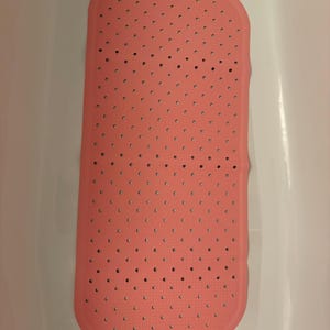 May include: A rectangular pink bath mat with rounded corners, featuring a grid of small circular drainage holes across the entire surface and a textured pattern of raised dots to provide grip on the floor of a white bathtub.