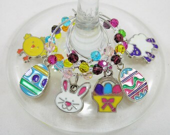 May include: Wine glass charms with Easter-themed pendants. Charms include a yellow chick, a white bunny, a white sheep, and colourful Easter eggs. The charms are attached to a beaded ring with colourful beads.
