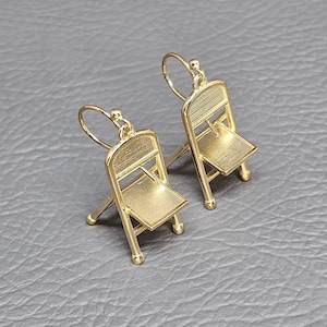May include: A pair of gold-tone earrings shaped like folding chairs. The earrings have a simple design and are a unique and stylish accessory.