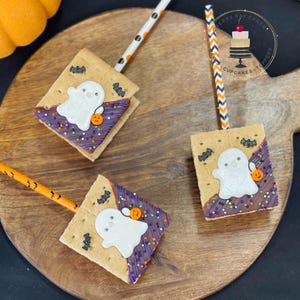 May include: Three graham cracker s'mores decorated with a ghost, bats, and sprinkles. The s'mores are on a wooden board with orange and white striped straws.