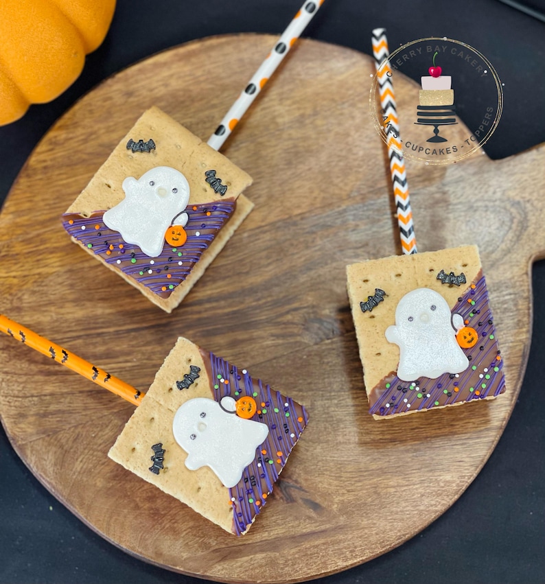 May include: Three graham cracker s'mores decorated with a ghost, bats, and sprinkles. The s'mores are on a wooden board with orange and white striped straws.
