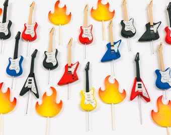 May include: Assortment of colorful electric guitar cake toppers, including red, blue, black, and yellow guitars, with some featuring flames. The toppers are on wooden sticks, ready to decorate a cake.