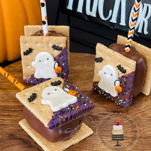 May include: Three ghost-themed s'mores pops with chocolate, graham crackers, and marshmallow topped with sprinkles, candy bats, and a white fondant ghost. The pops are on a wooden surface with a pumpkin and a sign that says "Trick or Treat".