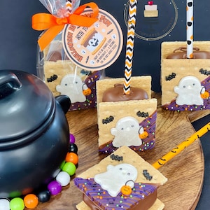 May include: A close-up of four Halloween-themed s'mores on sticks. Each s'more is made with graham crackers, chocolate, and a marshmallow topped with a white ghost and orange pumpkin. The s'mores are decorated with sprinkles and bats. The s'mores are on a wooden board with a black cauldron and candy in the background.
