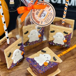 May include: A close-up of four ghost-themed s'mores on sticks. Each s'more is made with graham crackers, chocolate, and marshmallow, and is decorated with a white ghost, black bats, and purple and green sprinkles. The s'mores are on a wooden surface, and there is a tag that says "Halloween is with you" and a straw in each s'more.