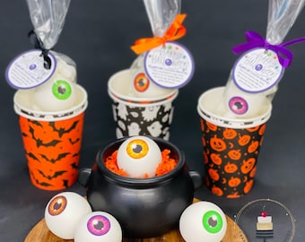 May include: Four white cups with Halloween themed designs, each containing a white eyeball bath bomb with a colorful iris. The cups are arranged around a black cauldron filled with orange shredded paper and a white eyeball bath bomb. The text on the cups reads "Happy Halloween".