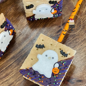 May include: A Halloween-themed graham cracker treat with a white ghost marshmallow, chocolate, and sprinkles. The treat is decorated with bats and a small orange pumpkin.