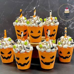 May include: Seven orange pumpkin-shaped cups with black jack-o'-lantern faces filled with white marshmallows, chocolate sauce, and sprinkles. The cups are arranged on a dark surface.