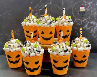 May include: Seven orange pumpkin-shaped cups with black jack-o'-lantern faces filled with white marshmallows, chocolate sauce, and sprinkles. The cups are arranged on a dark surface.