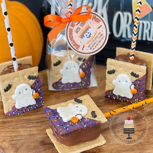 May include: Four chocolate-covered graham cracker s'mores with ghost-shaped marshmallows and Halloween sprinkles. The s'mores are on sticks and ready to be eaten. The text on the tag reads "Halloween is fun with you!"