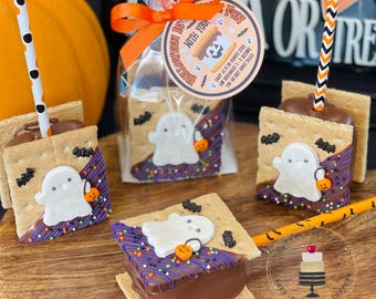 May include: Four chocolate-covered graham cracker s'mores with ghost-shaped marshmallows and Halloween sprinkles. The s'mores are on sticks and ready to be eaten. The text on the tag reads "Halloween is fun with you!"