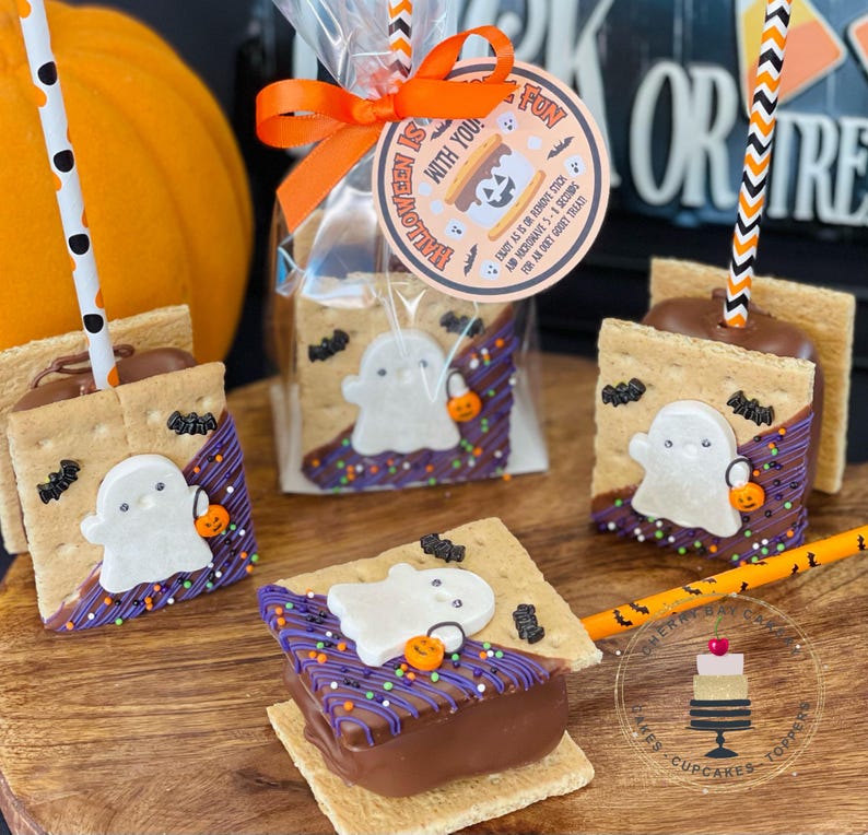 May include: Four chocolate-covered graham cracker s'mores with ghost-shaped marshmallows and Halloween sprinkles. The s'mores are on sticks and ready to be eaten. The text on the tag reads "Halloween is fun with you!"