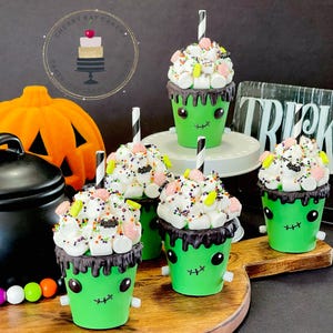 May include: Four green monster-themed hot chocolate cups with white marshmallow and candy toppings, black chocolate drizzle, and striped straws. The cups are on a wooden surface with a pumpkin and a black kettle in the background.