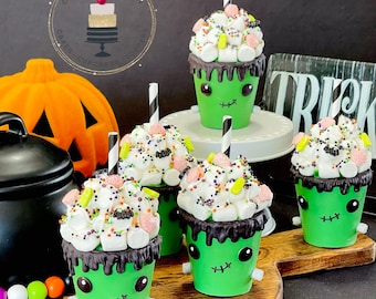 May include: Four green monster-themed hot chocolate cups with white marshmallow and candy toppings, black chocolate drizzle, and striped straws. The cups are on a wooden surface with a pumpkin and a black kettle in the background.