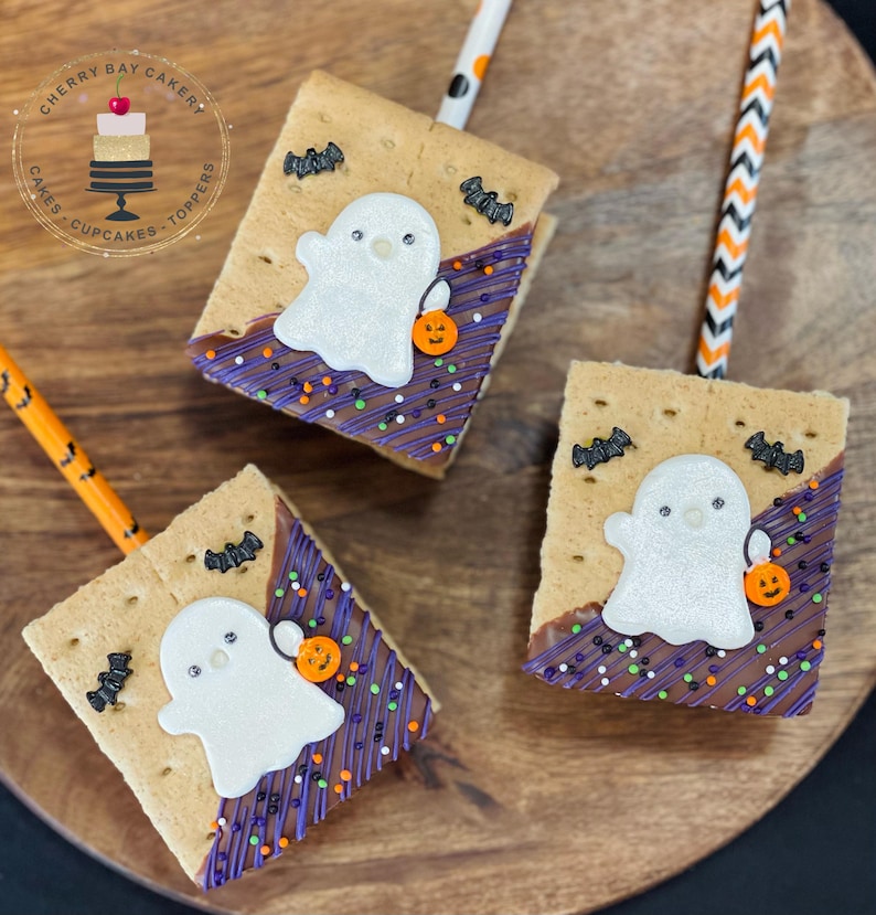 May include: Three graham cracker treats decorated with white ghost shapes, purple and brown icing, and sprinkles. The treats are on a wooden surface and each has a straw.
